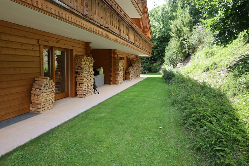 2-story wooden house with a covered balcony, stone pillars, and a well-maintained lawn surrounded by lush greenery