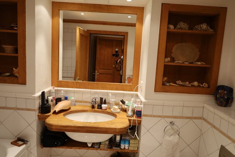 White tiled walls, wooden sink with countertop, mirror with wooden frame, wooden shelves on the wall, toilet visible, towel rack, and various toiletries