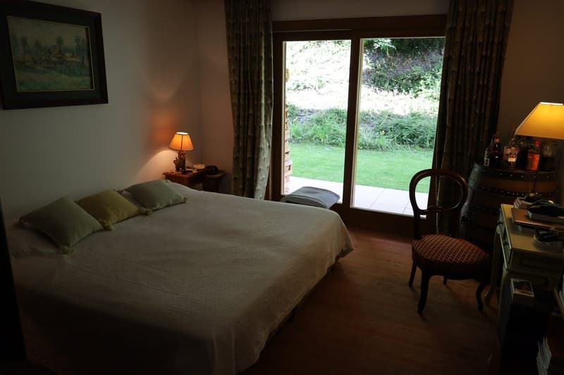 Spacious bedroom with large windows overlooking a lush green garden, hardwood floor, a queen-size bed with green bedding, a side table with a lamp, and a chair in the corner.