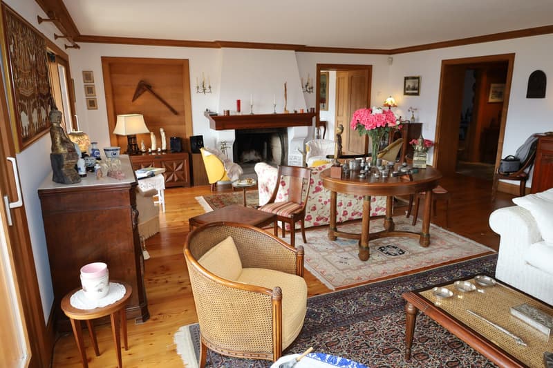 Cozy living room with a fireplace, wooden furniture, floral accents, and a round dining table. The room features a mix of traditional and eclectic decor elements.
