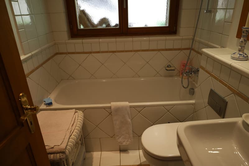 The image shows a bathroom with a tiled floor and walls, a bathtub, a toilet, and a window with a frosted glass pane.