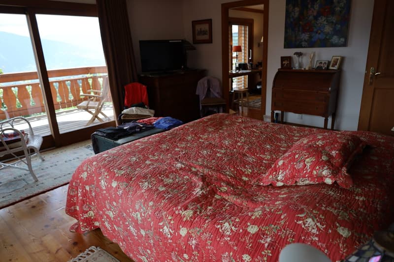 Spacious bedroom with a large floral-patterned comforter on the bed, wooden furniture including a dresser and shelves, and a sliding glass door leading to a balcony or terrace with a view.