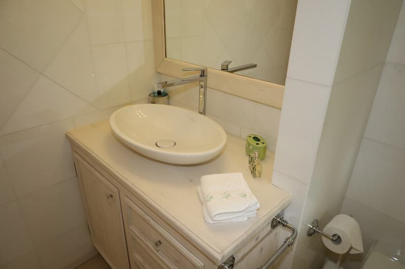 tiled wall, mirror, white ceramic sink, wooden cabinets, stacked towels, roll of toilet paper, tissue container, soap dispenser