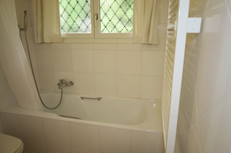 Bathtub, handheld showerhead, window with white curtains, white tiled walls, toilet