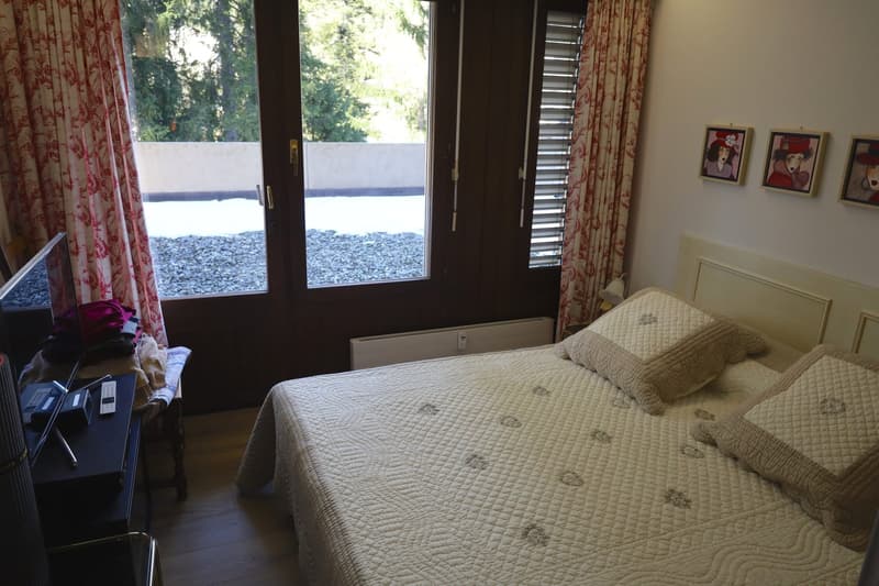 A double bed with beige comforter, a desk, a television, and a chair. Windows with curtains, pictures, a white heater on the wall.