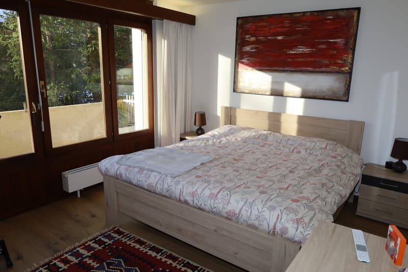 large double bed with floral bedding, two nightstands with lamps, large picture frame, wooden flooring, large glass doors leading to balcony, radiator, rug, remote