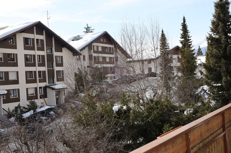 Modern apartment building, multiple units, brown and white walls, several parked cars, snow on roofs, bare trees, mountains in background