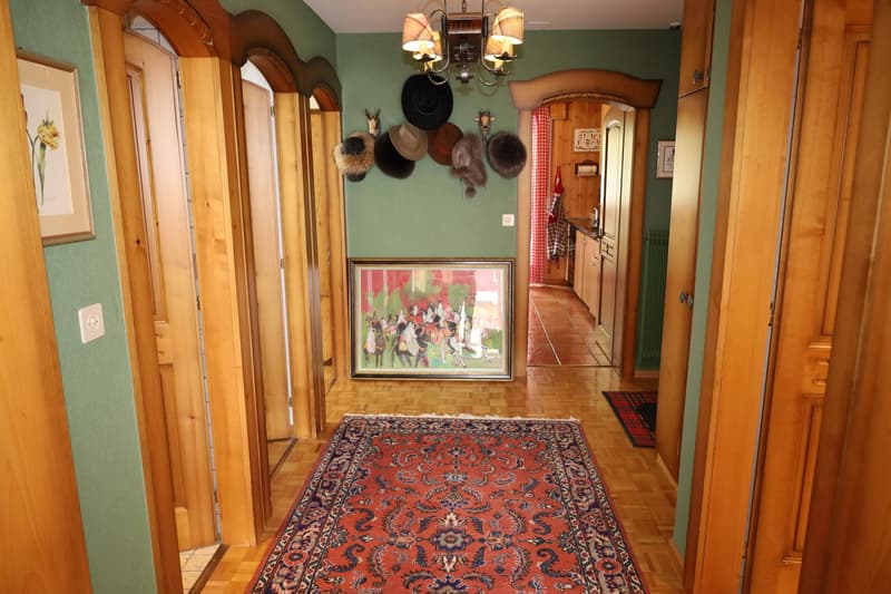 A hallway with wooden doors and green walls, oriental rug, picture frame on the wall, hats and other items on the wall