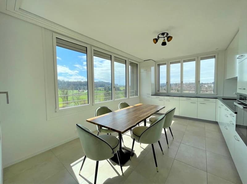 Spacious open-plan kitchen and dining area with large windows offering a scenic view of the surrounding countryside. The room features a wooden dining table with chairs, modern white cabinets, and tiled flooring. The kitchen appears well-equipped with app