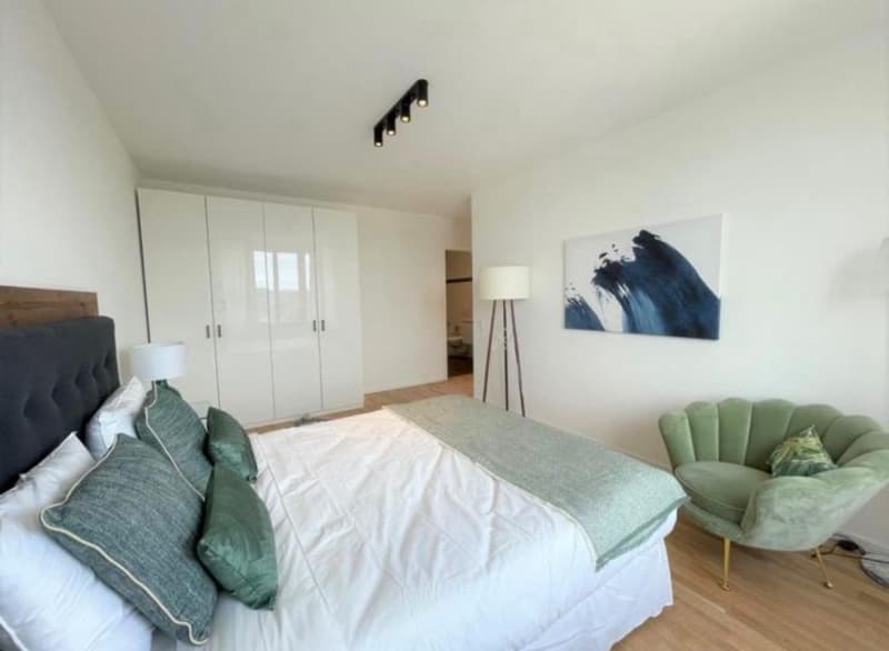 Spacious bedroom with white walls, hardwood floors, large wardrobe, king-size bed with green and white bedding, green armchair, and a large abstract painting on the wall.