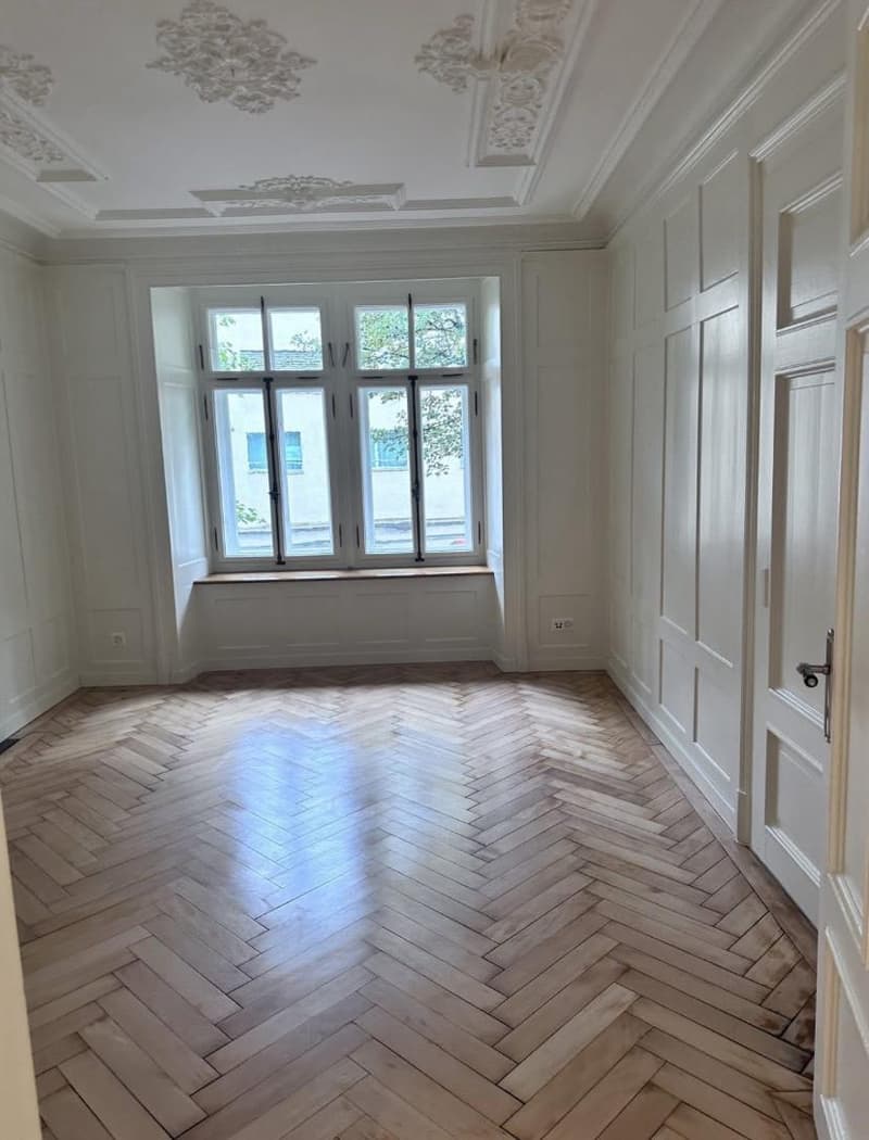 Empty room, herringbone wooden floor, white walls, large window with bench, ornate ceiling