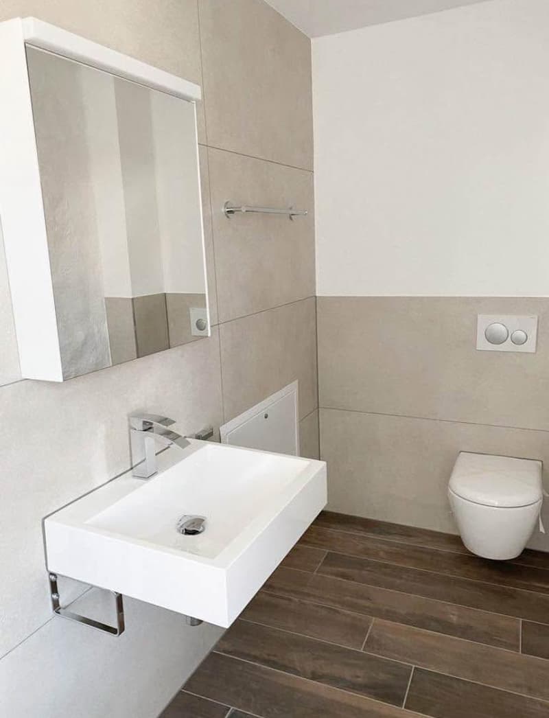 Modern bathroom, white sink, wall-mounted mirror, wooden flooring, wall-mounted toilet, and toilet controls