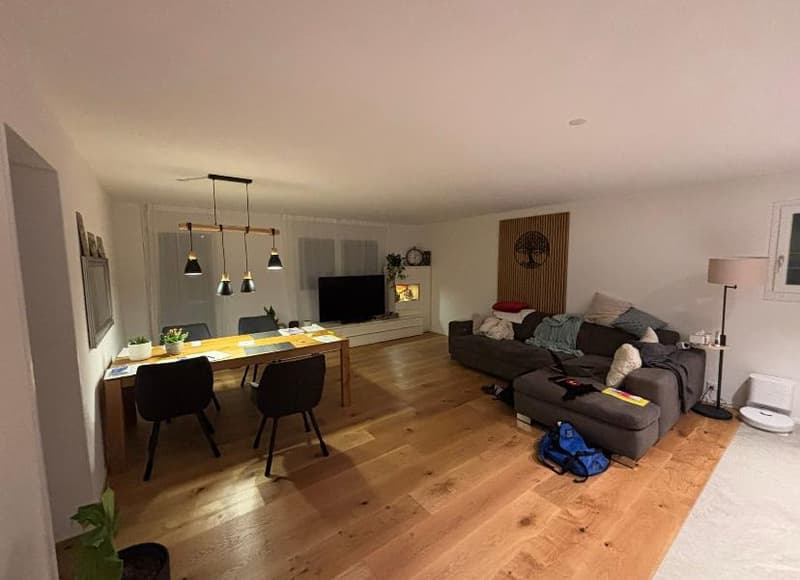 Living room with wooden floor, open to dining area, couch, dining table with chairs, mounted TV, white ceiling, large windows with curtains, ceiling lamps, side table, decorative objects