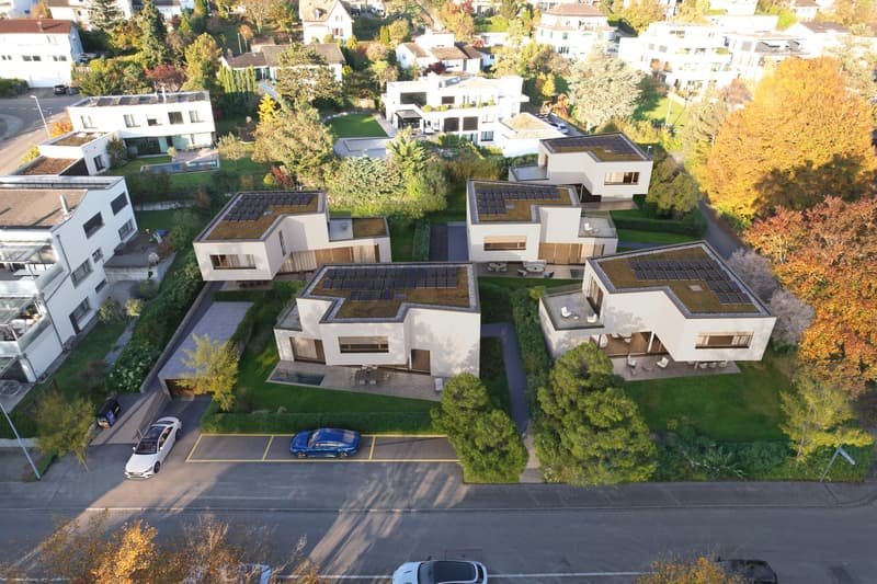 Cluster of contemporary houses, with solar panels on the roofs, driveways, parking areas, surrounded by greenery.