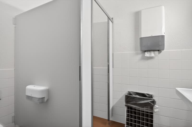 Bathroom stall, white walls, white tile floor, white door, toilet paper holder, waste basket, glass door, sink
