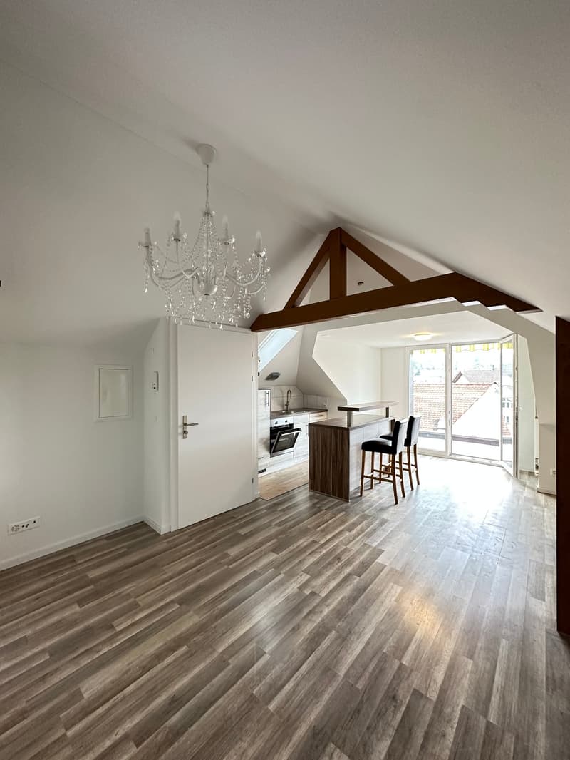 Open-concept living space with hardwood flooring, white walls, exposed wooden beams, a crystal chandelier, and a kitchen area with a counter and bar stools. There is a sliding glass door leading to a balcony or terrace.