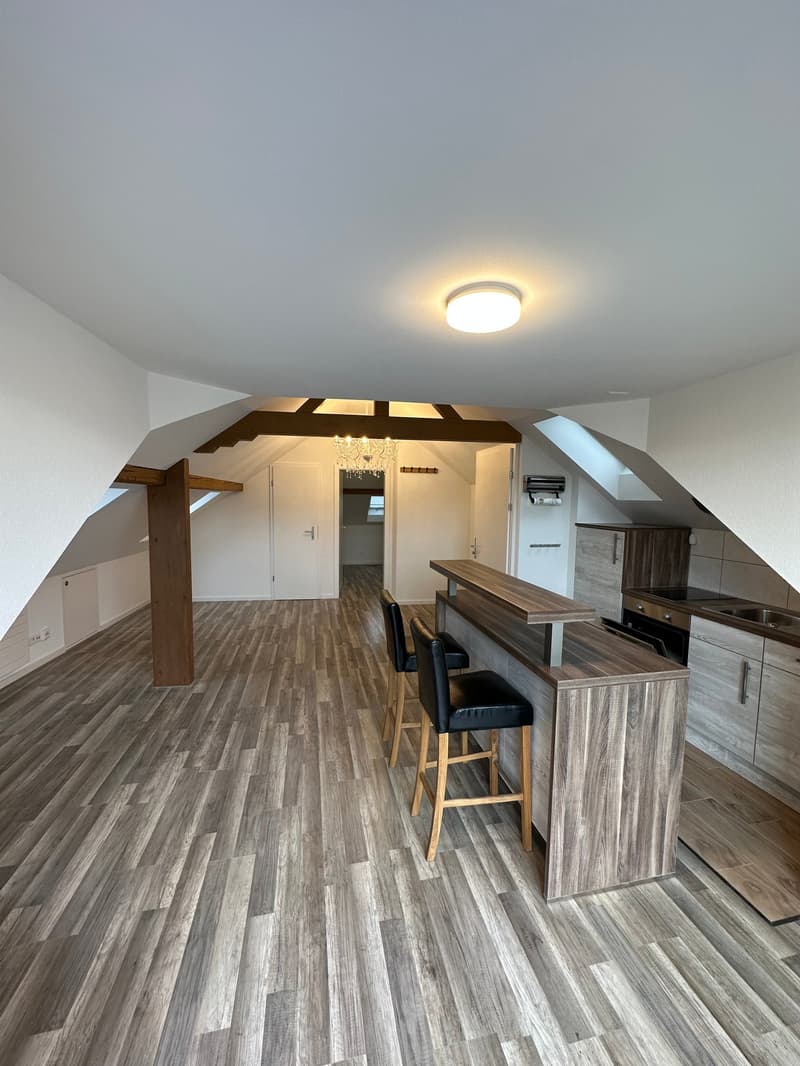 Open-concept living space with hardwood-style flooring, exposed wooden beams, a kitchen counter with bar stools, and a chandelier. The space appears to have a balcony or terrace and includes a dishwasher in the kitchen area.