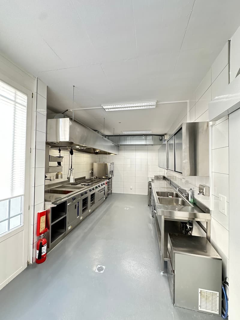 Modern commercial kitchen with stainless steel appliances, gas stoves, ovens, sinks, fire extinguisher, tiled floor, white walls, and ceiling lighting