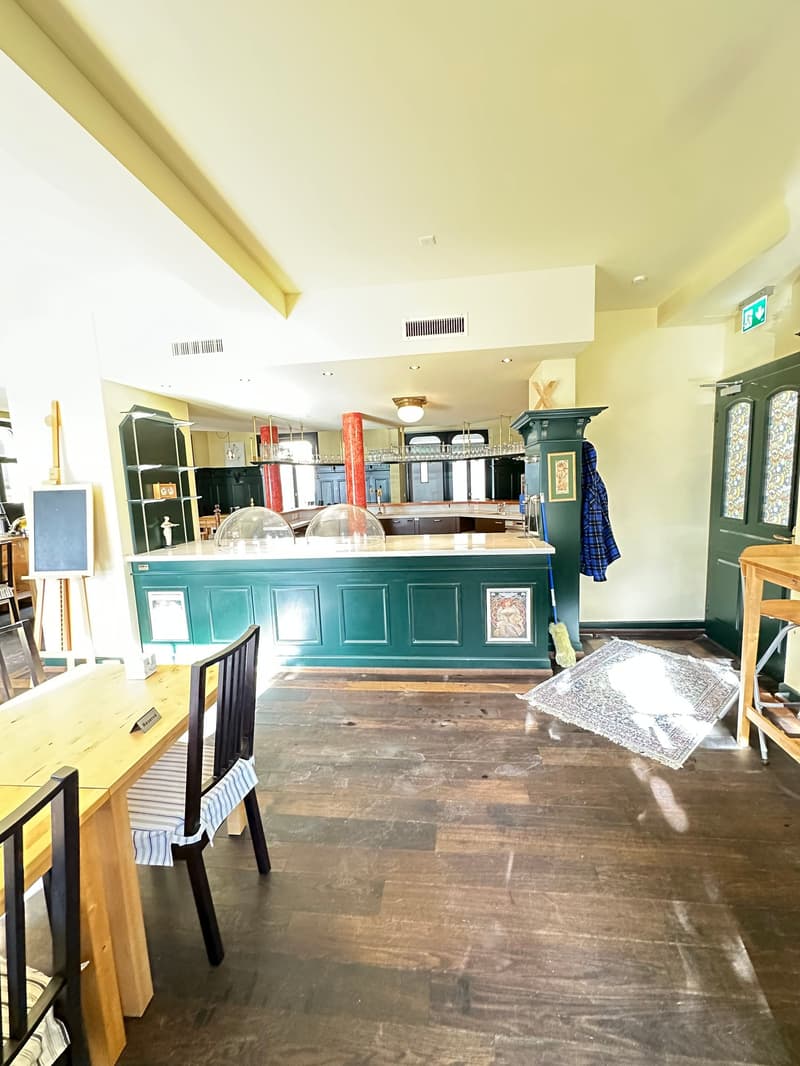 A green and white counter is placed in the middle. Wooden tables and chairs are beside the counter. Shelves are on the left side. There are two glass domes above the counter. A blackboard with a stand is on the left. A coat is hanging on the post near the
