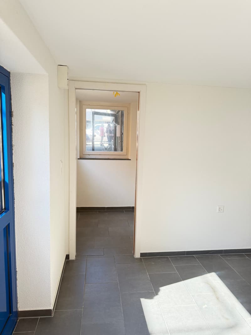 Empty room, white walls, gray tiles, blue door, window with view to outside