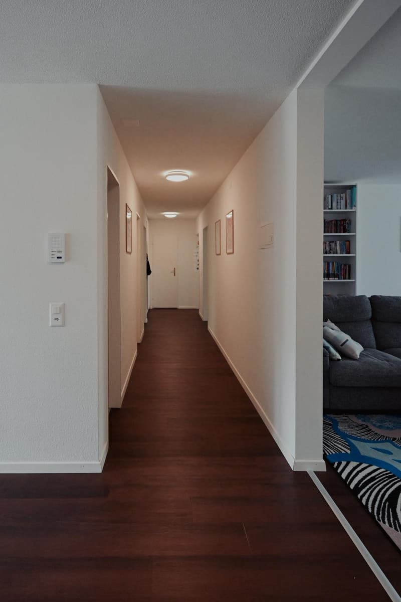 long hallway, white walls, wooden floors, mounted lights, closed door, white pillar