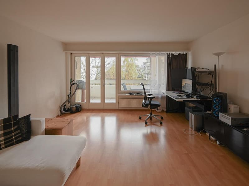 A room with a desk and chair, an office setup, a couch, wooden floor, and a balcony with glass doors
