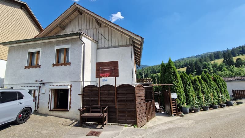 2-story house with wooden siding, white exterior walls, and a basketball hoop. There are balconies on the second floor and a parking space in front of the house. The house is surrounded by trees and greenery in a mountainous area.