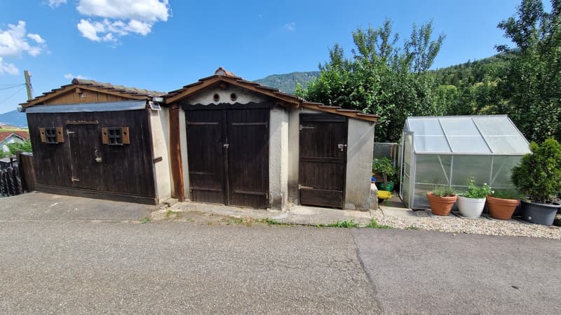 The image shows a group of three wooden outbuildings, likely sheds or storage structures, with a greenhouse structure visible next to them. The buildings have a rustic, traditional appearance with wooden doors and windows. The surrounding landscape includ
