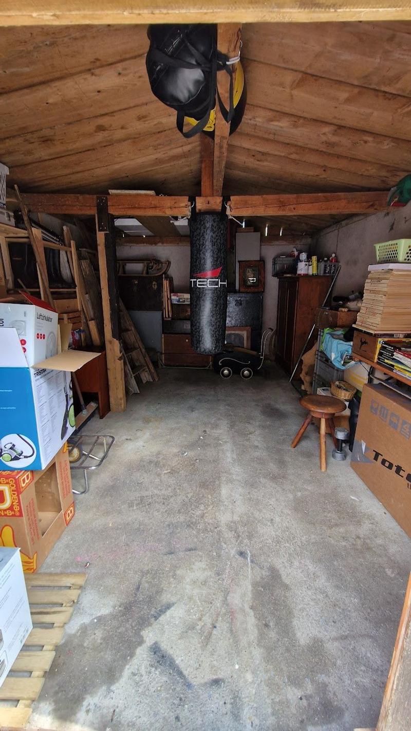 The image shows the interior of a garage or storage space. The space contains various items such as boxes, tools, and a punching bag. The ceiling is made of wooden beams, and the floor is concrete.