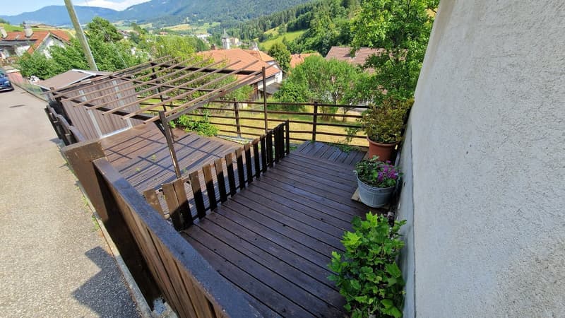 Wooden balcony with railing, overlooking a scenic mountain landscape with houses and greenery