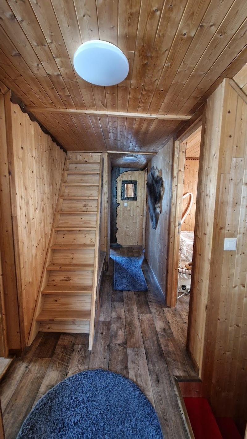 The image shows the interior of a wooden cabin or tiny home. The space features a wooden ceiling and walls, with a staircase leading to an upper level. The floor is covered with a blue rug, and there is a light fixture hanging from the ceiling.