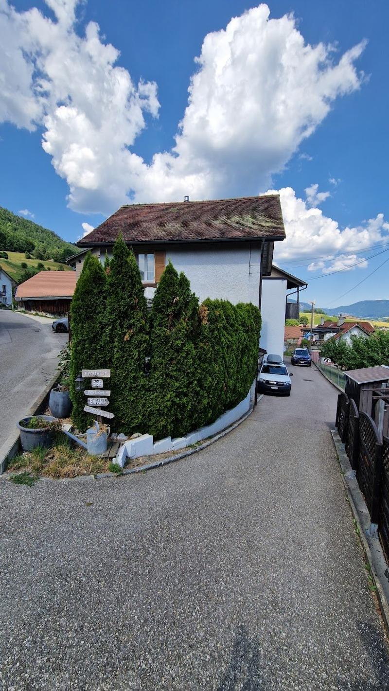 Two-story house with a tiled roof, surrounded by lush greenery and trees. The house has a paved driveway leading to it, and there appears to be a balcony or terrace on the upper level.