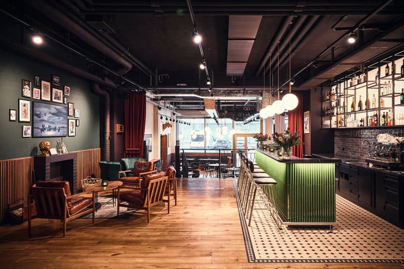 The image shows a cozy and stylish restaurant or bar interior. The space features a mix of vintage and industrial elements, with exposed brick walls, wooden floors, and a green-painted bar counter. There are comfortable leather armchairs and sofas, as wel