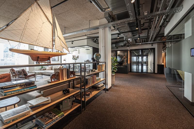 Open concept office space with high ceilings, exposed ductwork, and wooden shelving units. The space features a large sailboat model hanging from the ceiling, as well as various decorative items and books on the shelves. The office has a modern, industria