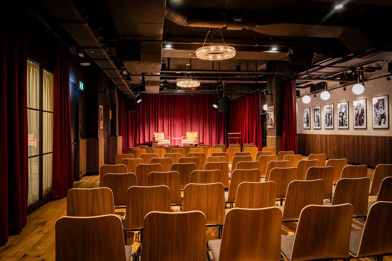 A small theater or performance space with rows of wooden chairs facing a stage with red curtains. The room has a high ceiling with exposed pipes and ductwork, and several chandeliers hanging from the ceiling. The walls are decorated with framed black and