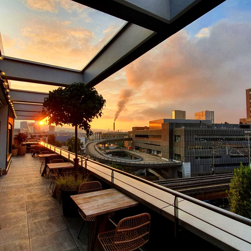 Outdoor balcony or terrace overlooking a city skyline at sunset. The balcony has wooden furniture and a potted plant, with a panoramic view of the cityscape including high-rise buildings and a smokestack in the distance.