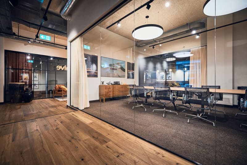 Large open office space with hardwood floors, exposed ceiling beams, and modern furniture. The space includes a conference table with chairs, a wooden credenza, and artwork on the walls. There is also a fireplace visible in the background.