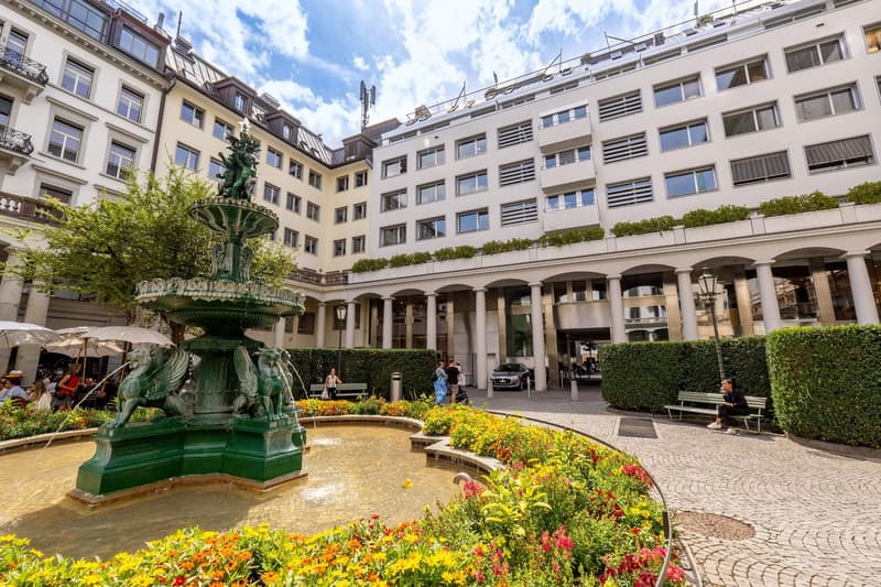 white apartment building, green fountain, large bushes, people sitting on benches, street lamp, parked car, white umbrellas