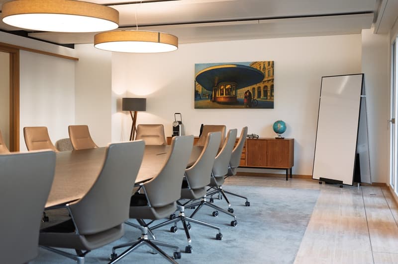 spacious room, white walls, large wooden table, multiple modern chairs, lamp, wooden desk, painting, whiteboard, rug