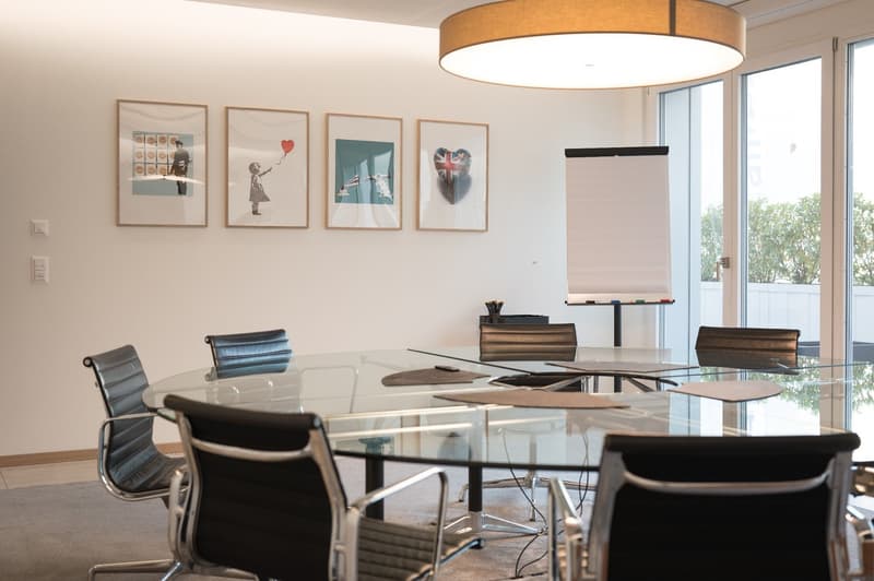 Meeting room with glass table, black leather chairs, glass doors, roll-up board, framed pictures, beige lighting, and carpet floor.