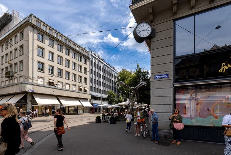 People walking, outdoor cafe, buildings, clock on wall, street light, plants, and trees