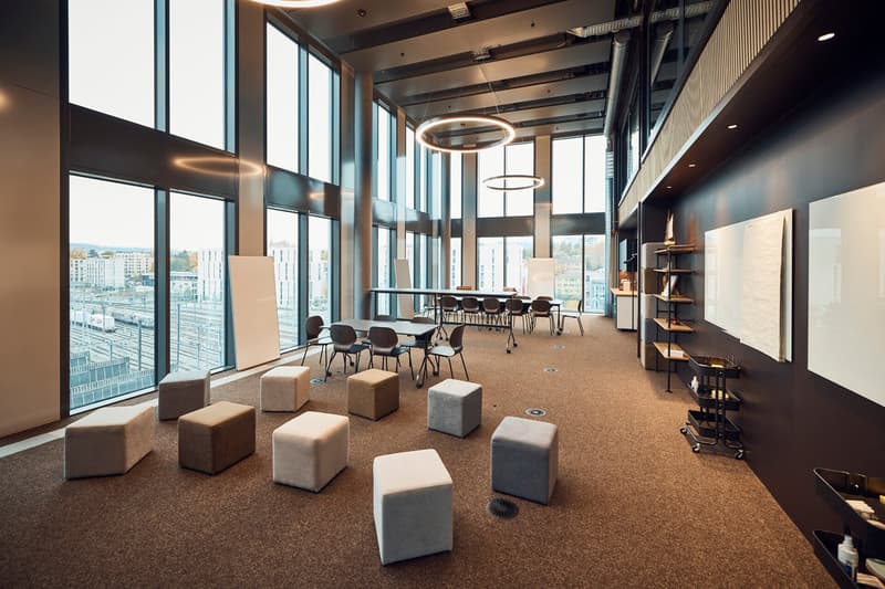 A large open room with carpeted floor, several tables, chairs, and circular ottomans, large windows with city view