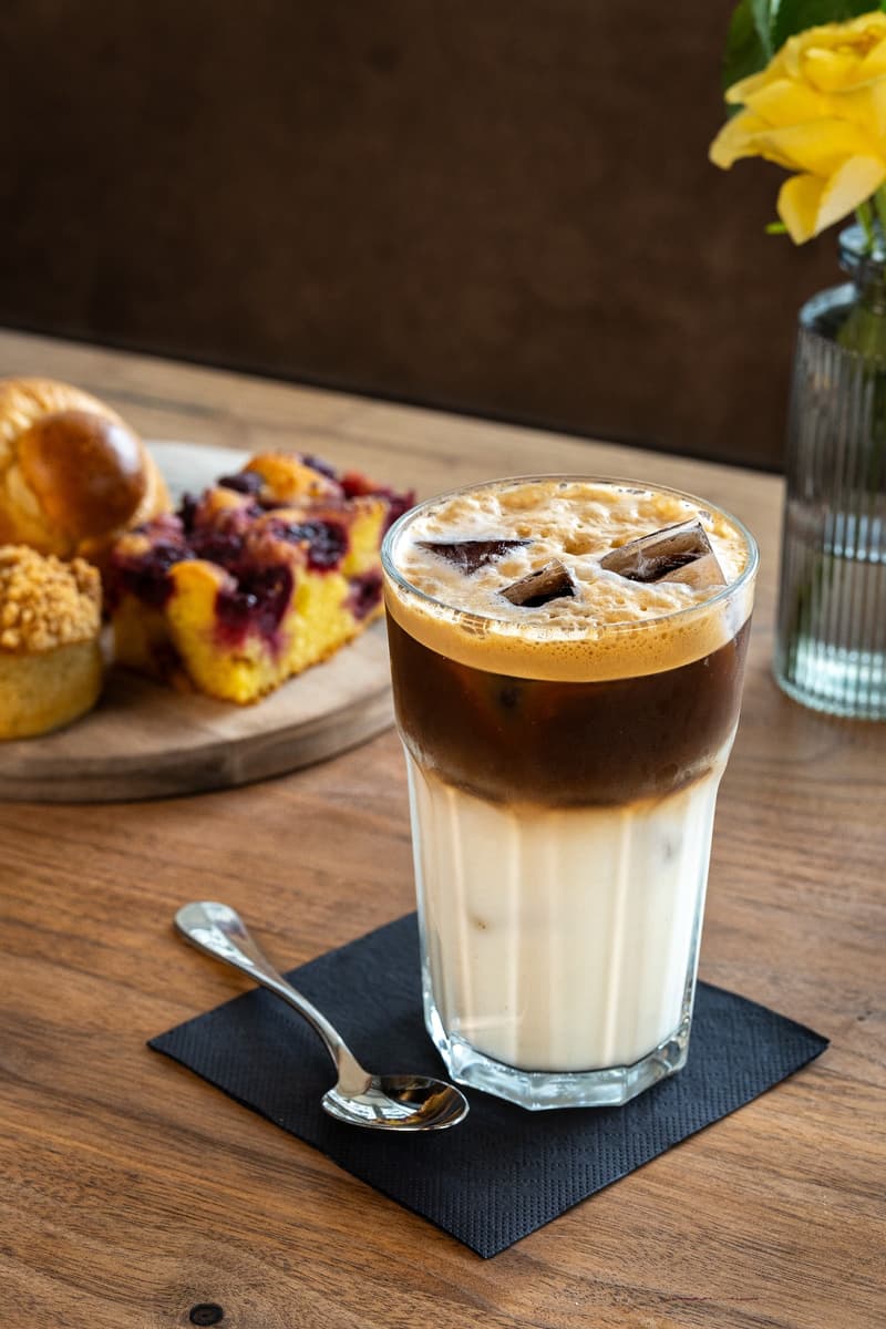 Cold brew coffee with milk, topped with ice cubes, served in a tall glass on a wooden table with food on a plate and a spoon