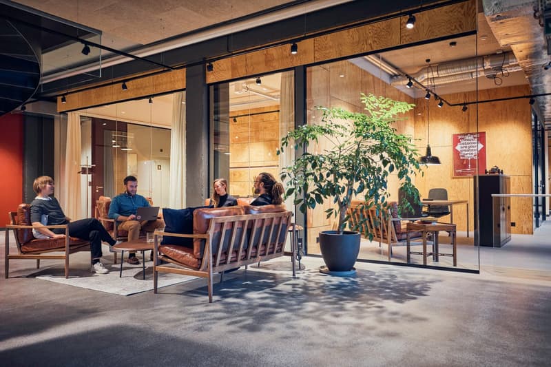 Open concept office with modern furniture and decor. Glass partitions, wooden floors, greenery, and large windows. Glass walls and a staircase visible.