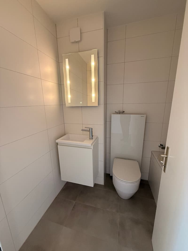 Modern bathroom with white walls, gray tiled floor, white vanity with sink, a mirror with integrated lighting, a modern toilet, and a bathtub