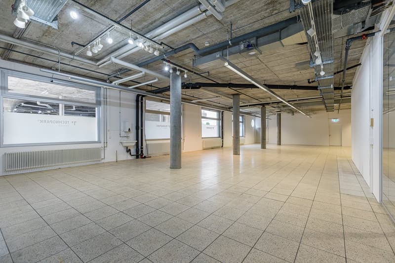 Large open commercial space with concrete ceiling, exposed pipes and ductwork, tile flooring, and windows along one wall. The space appears to be a warehouse or office area with a projector screen visible.