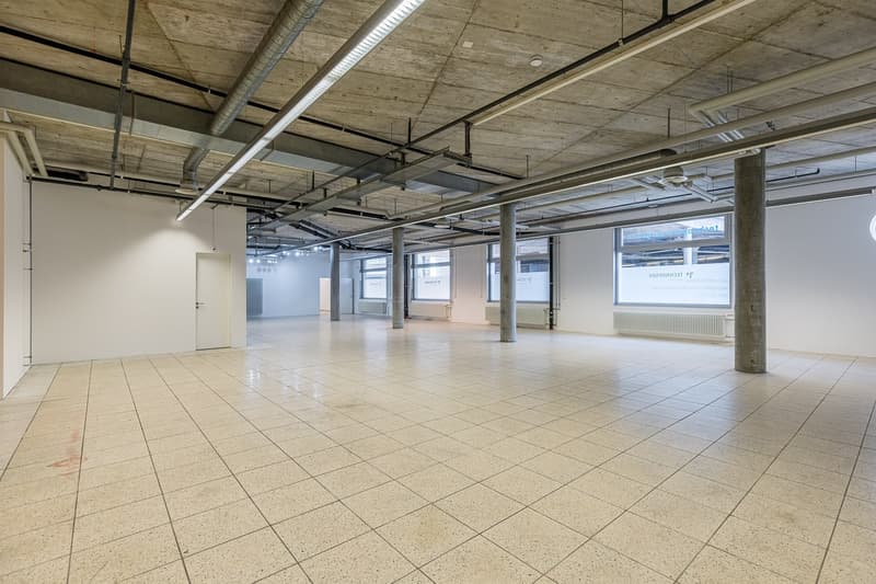 Large open-plan office space with concrete ceiling, exposed ductwork, and tiled flooring. The space appears to be a commercial or industrial building that has been converted into an office or workspace. There are several windows along the walls providing