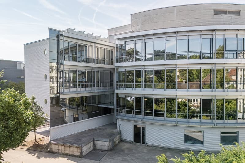 Multi-story modern office building with glass walls and balconies, surrounded by trees and greenery