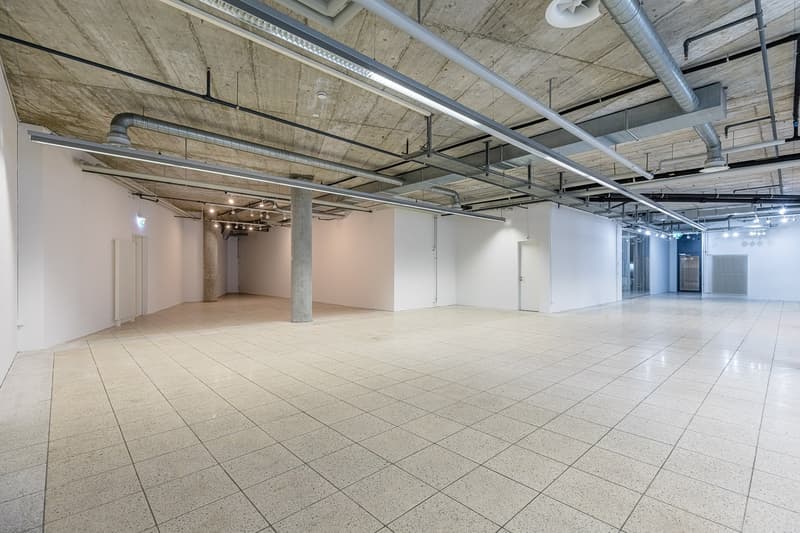 Large open commercial space with exposed concrete ceiling, ductwork, and lighting. The space has a tiled floor and appears to be a warehouse or industrial-style office area.