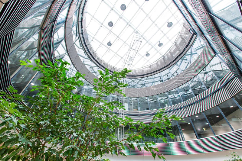 Large glass dome-shaped atrium with a curved, geometric glass and metal structure. The atrium features lush greenery, including several potted plants and trees, creating a natural, indoor garden-like environment.